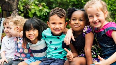 A group of kindergarten aged children sit in a row outdoors with their arms around each other smiling.